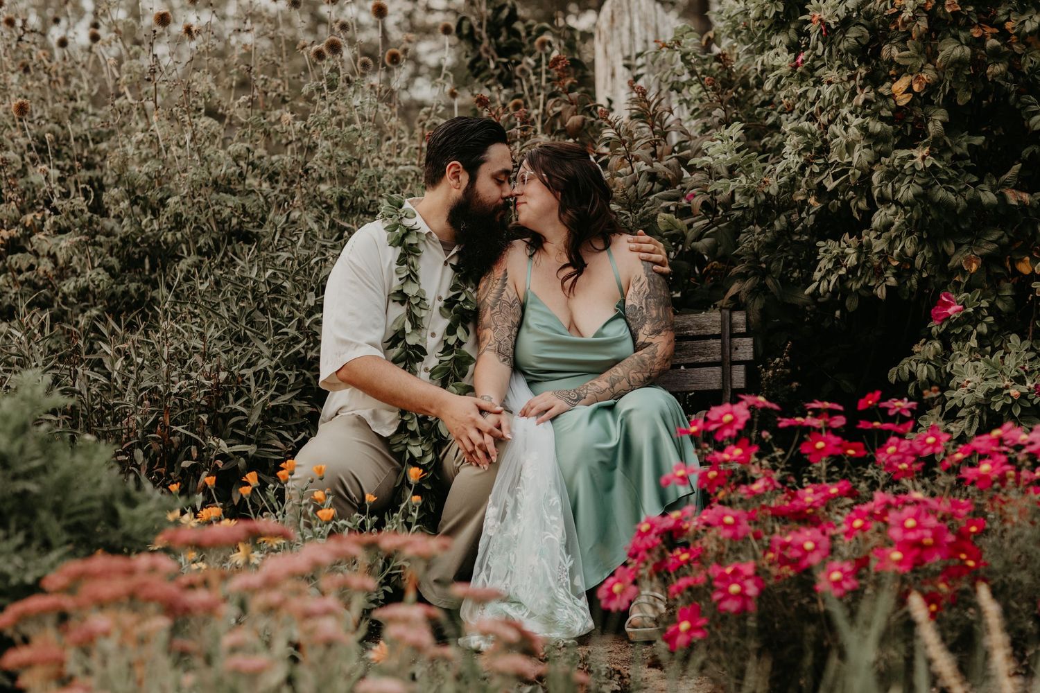A romantic scene in a garden with vibrant pink flowers, green foliage, and traditional Hawaiian leis around their necks.