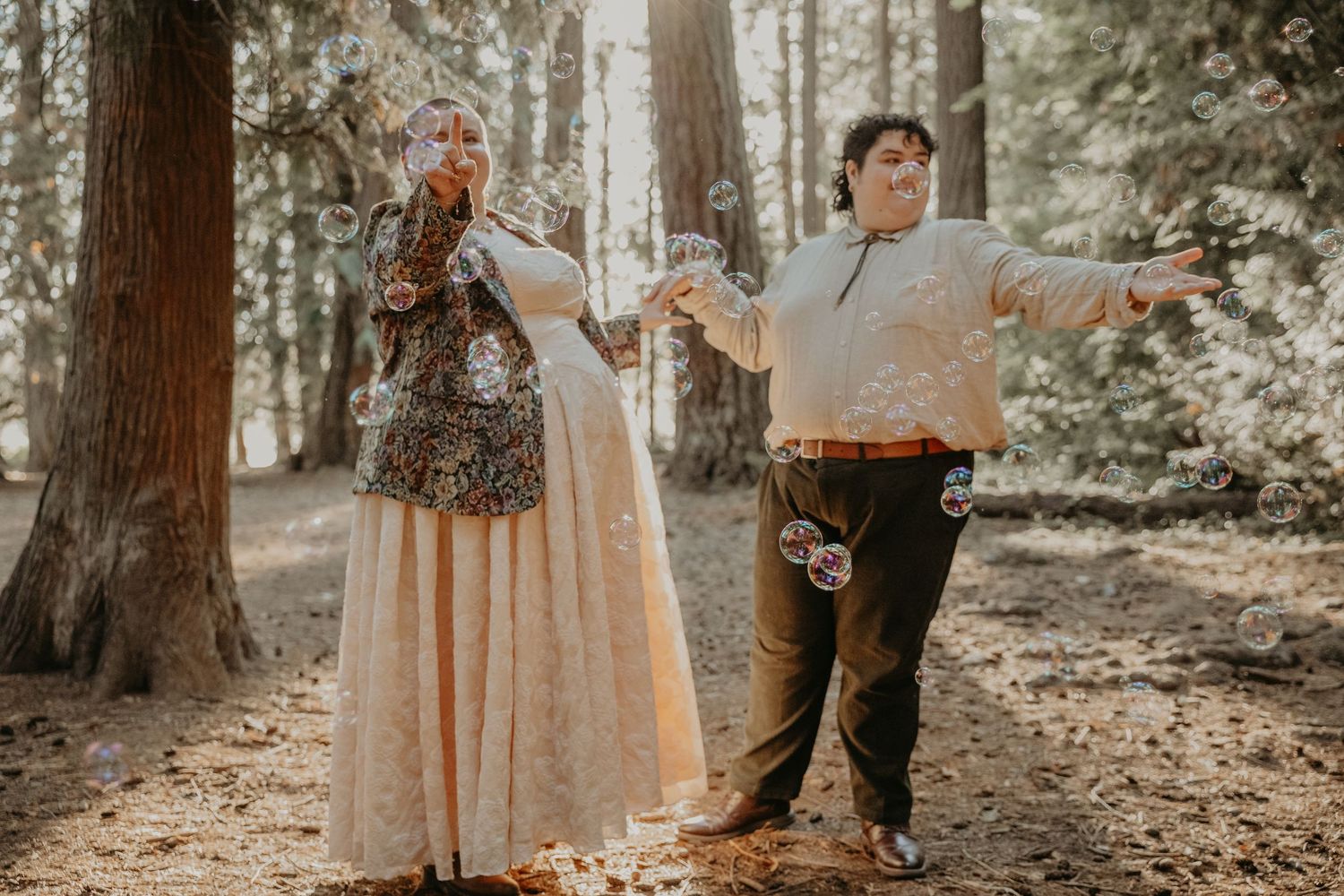 A couple poses playfully with outstretched arms in a sunlit forest setting.