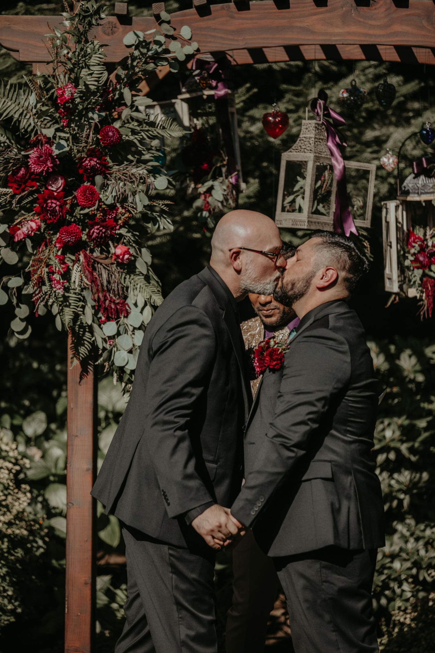 A romantic moment captured under a wooden pergola decorated with vibrant red flowers in a garden setting.