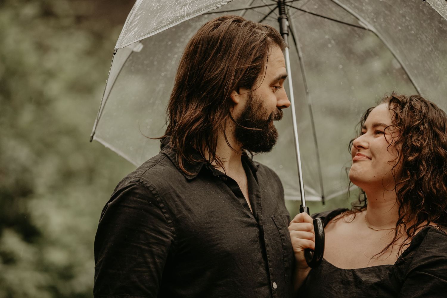 Romantic couple sharing tender moment under clear umbrella in moody forest setting.
