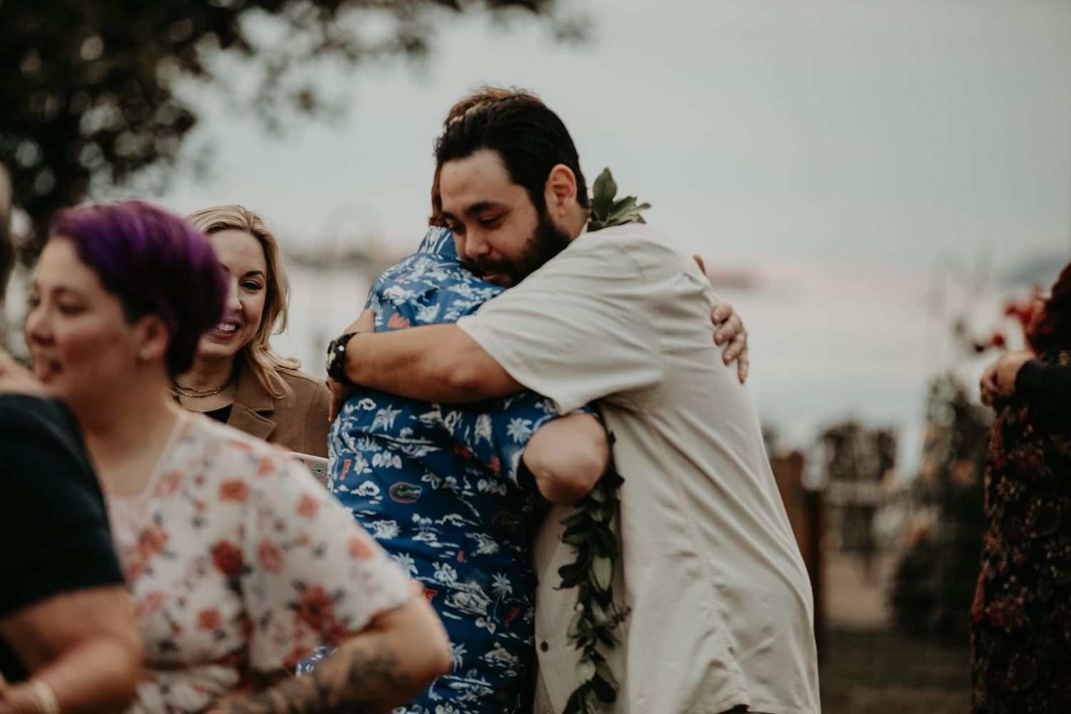 Two people share a warm embrace while others gather in the background at an outdoor event.