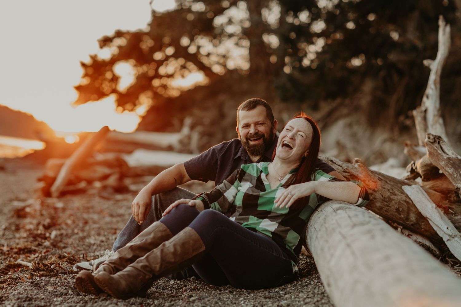A joyful couple laughs while sitting on driftwood at sunset by the beach.