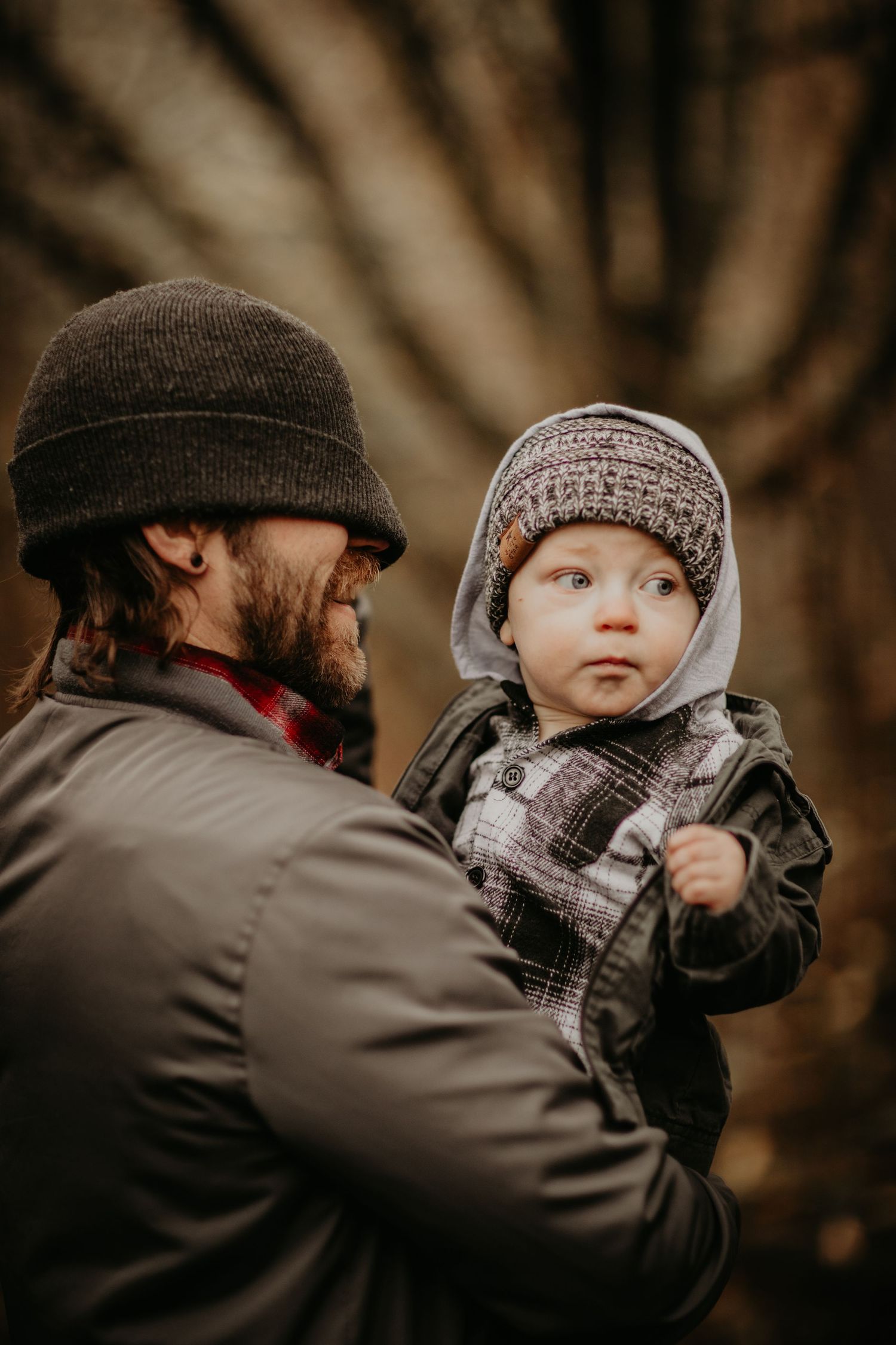 A parent wearing a brown beanie holds a small child bundled in winter clothing during an autumn outdoor photo session.