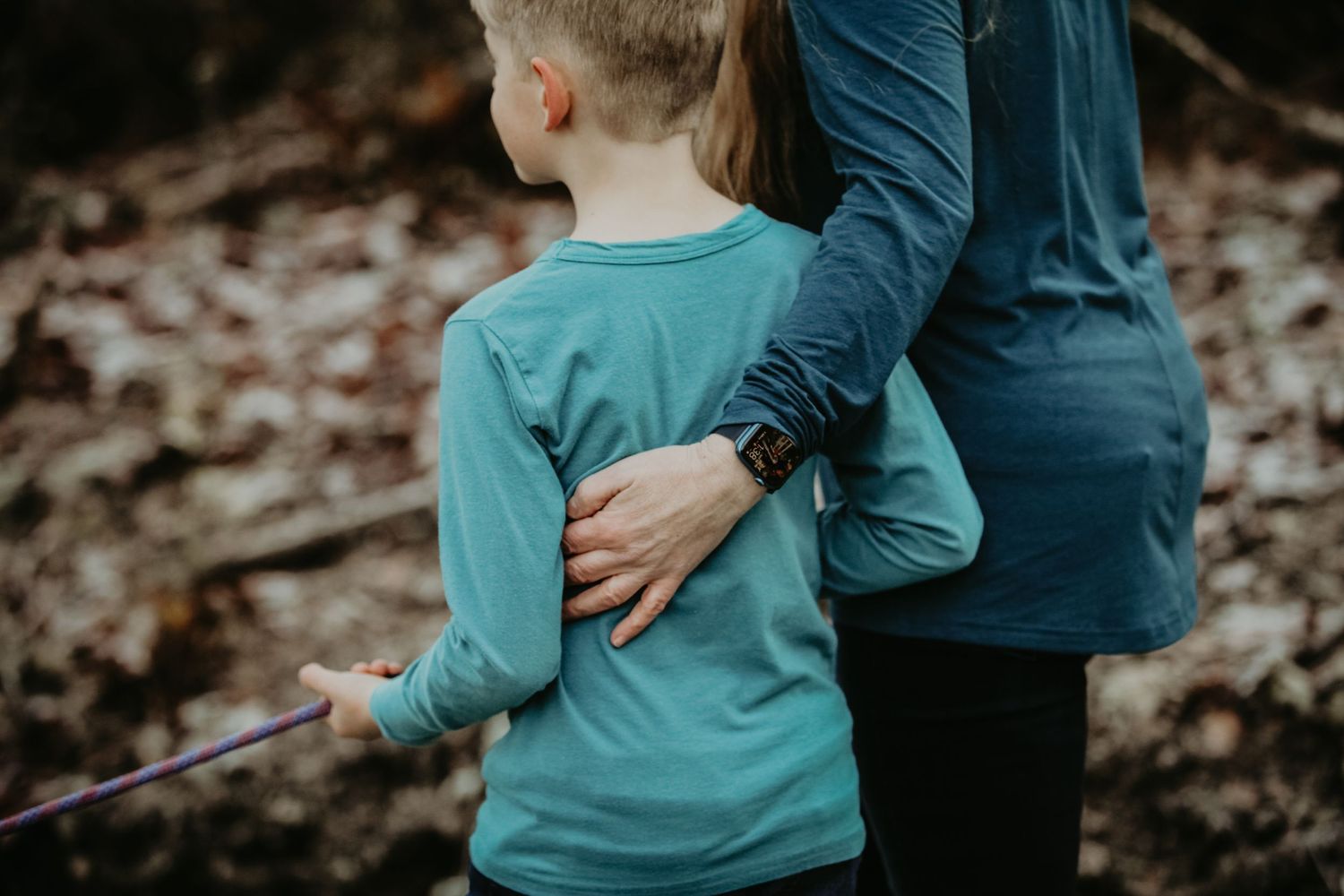 A caring embrace between parent and child wearing teal and navy blue shirts standing in an autumn forest.