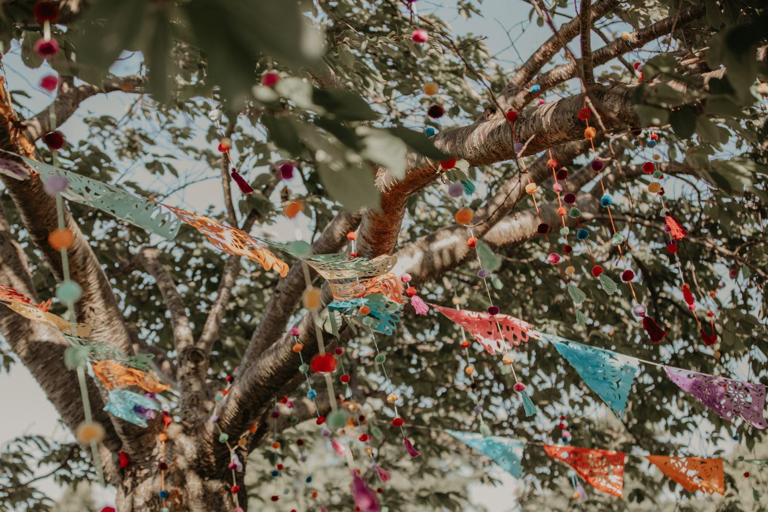 A wedding ceremony setup in a garden with white chairs and colorful floral decorations hanging from tree branches.