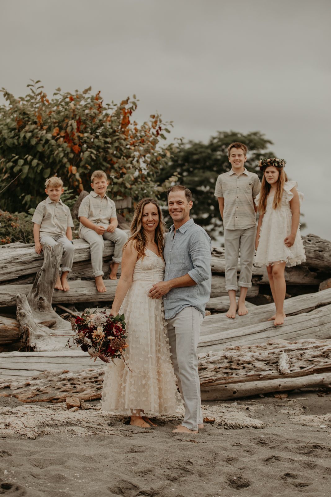 A beach wedding party poses on driftwood logs against an overcast sky.