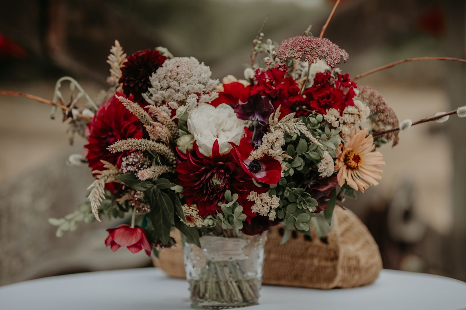 Elegant floral arrangements in deep red and white tones displayed in rustic vases and baskets at an outdoor event.