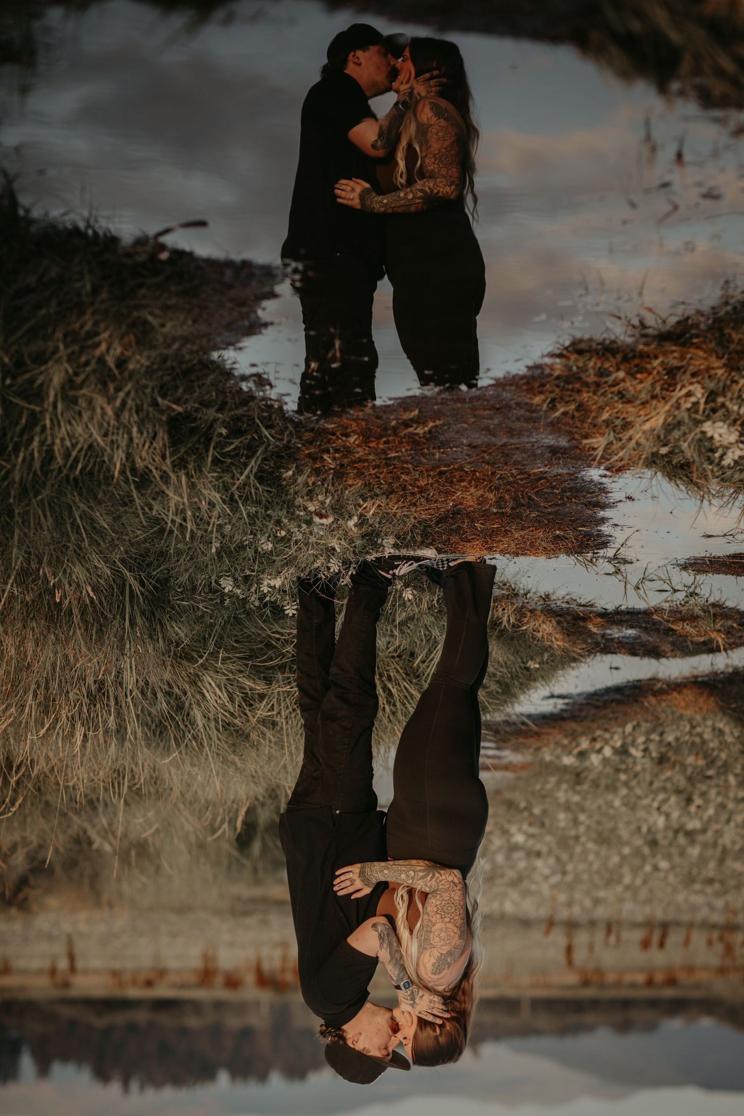 Couple embraces on a path with their reflection mirrored in water below during golden hour sunset.