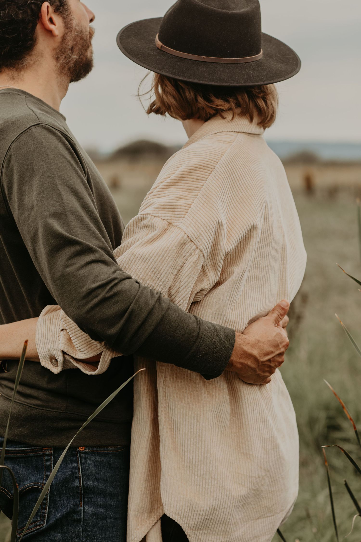 A couple embraces in a rustic outdoor setting, with person wearing a brown fedora and beige sweater against a serene landscape.