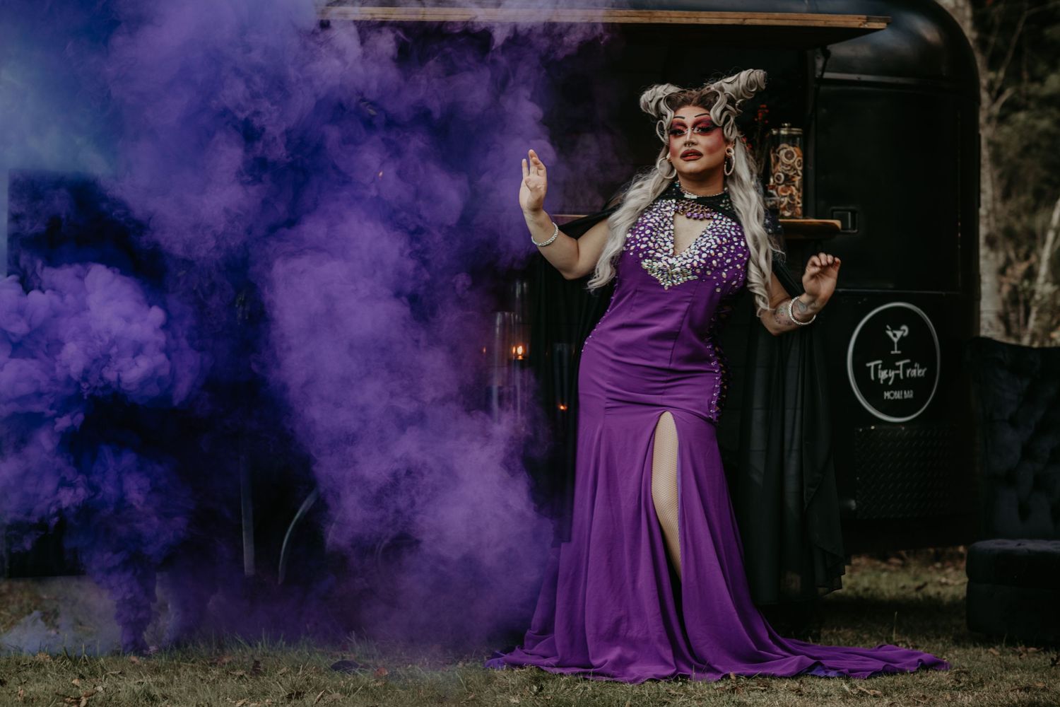 A person in a purple dress stands amid purple smoke while striking a dramatic pose against a dark backdrop.