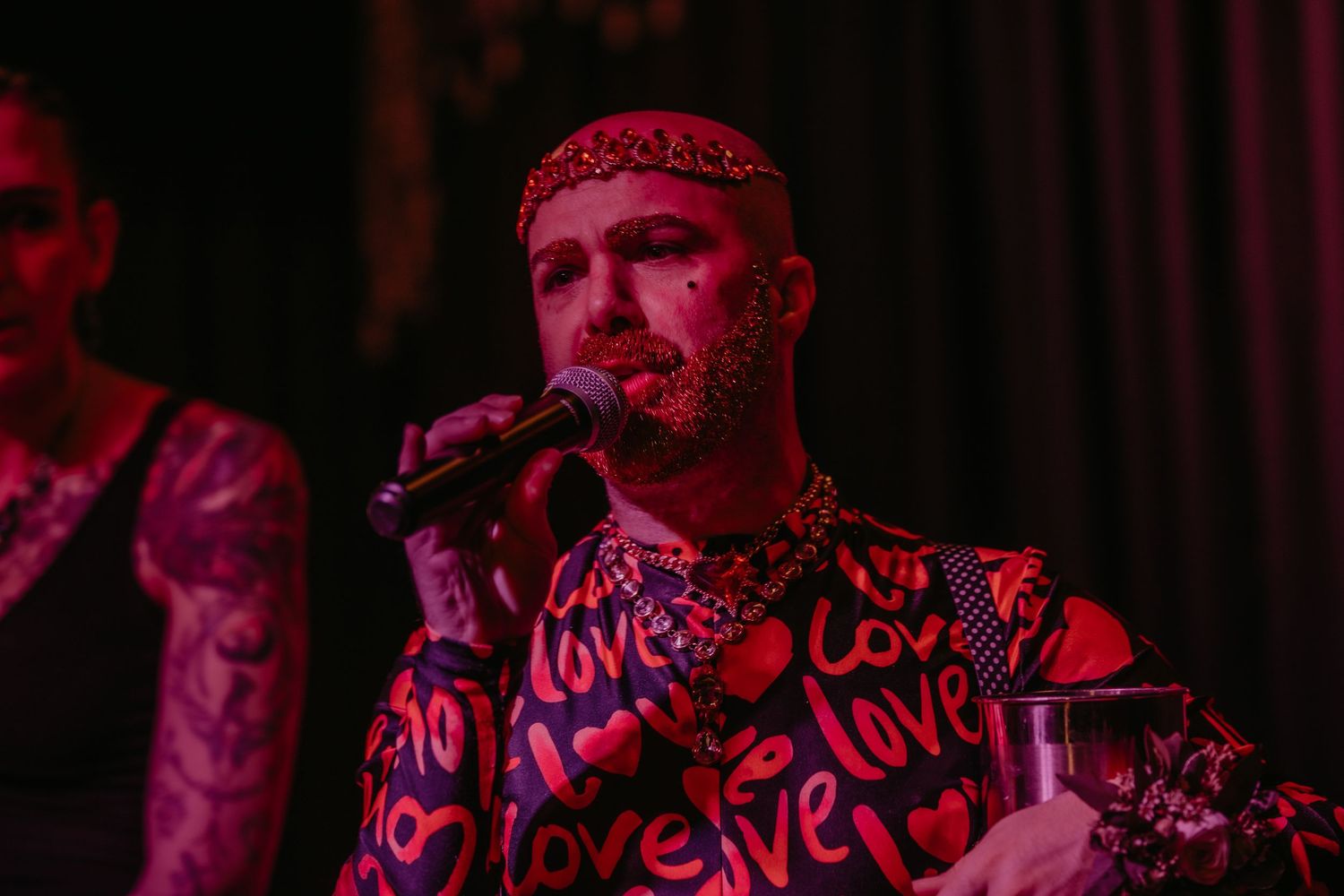 A performer in a red and black 'Love' patterned shirt sings into a microphone on stage under dramatic lighting.