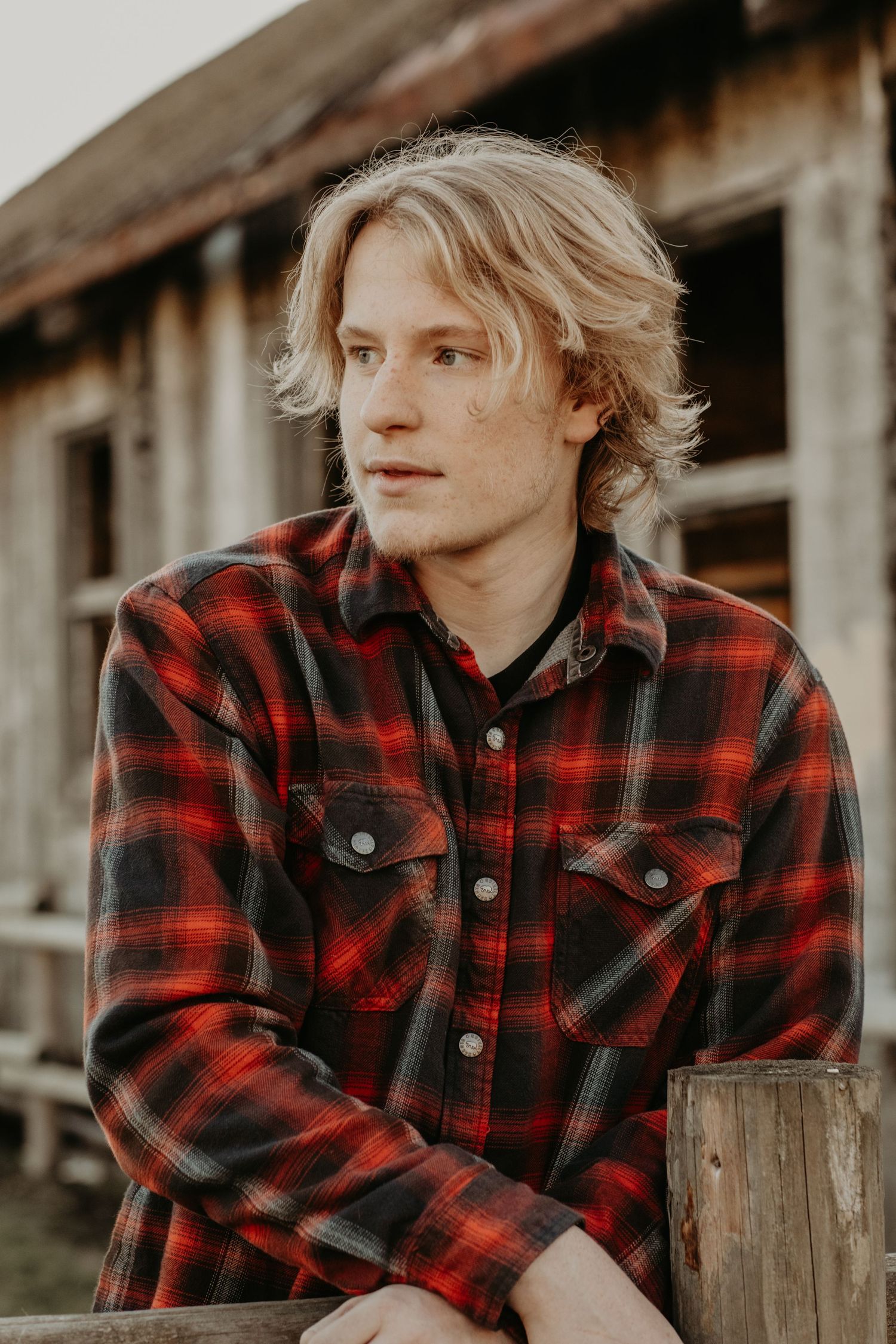 Person in red and black plaid flannel shirt looking pensively to the side against rustic wooden building exterior.