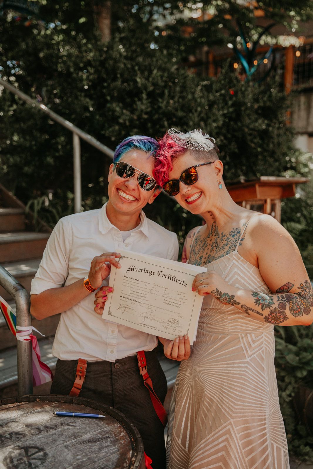 A couple in sunglasses and light-colored attire share joyful moments while holding a marriage certificate outdoors.