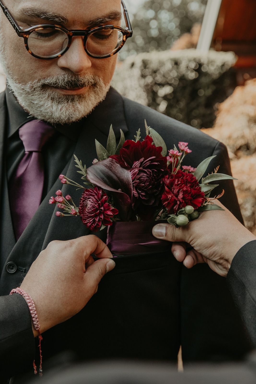 Close-up sequence showing someone in glasses pinning a deep burgundy boutonniere to a dark suit jacket.