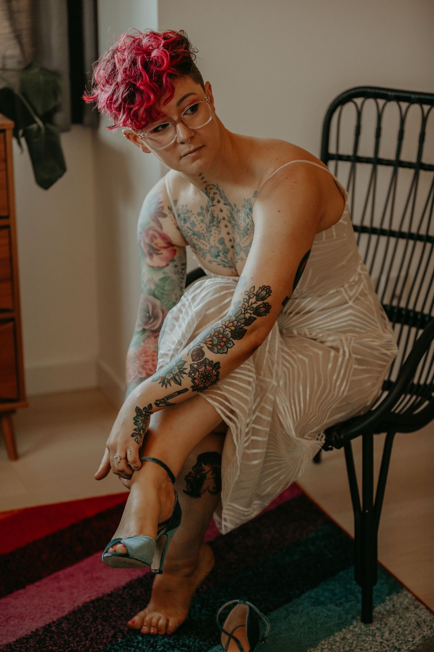 A person with pink hair and tattoos sits on a rattan chair wearing a white dress.