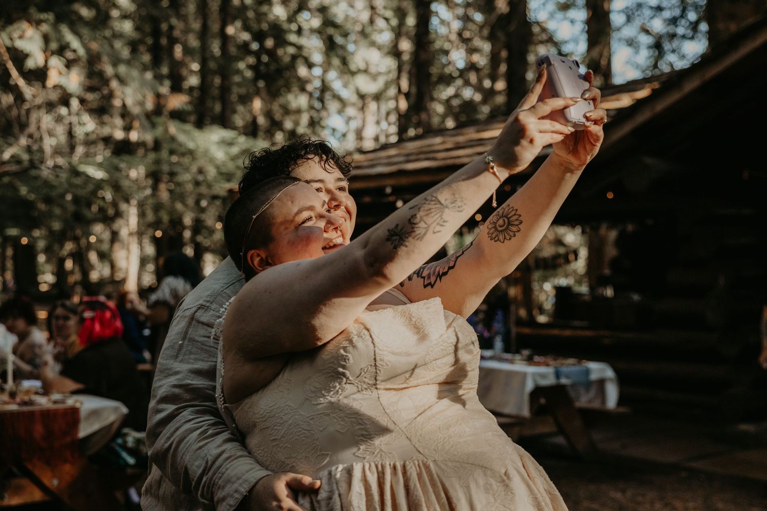 A group of wedding guests raise their glasses in celebration during an outdoor reception in a wooded setting.