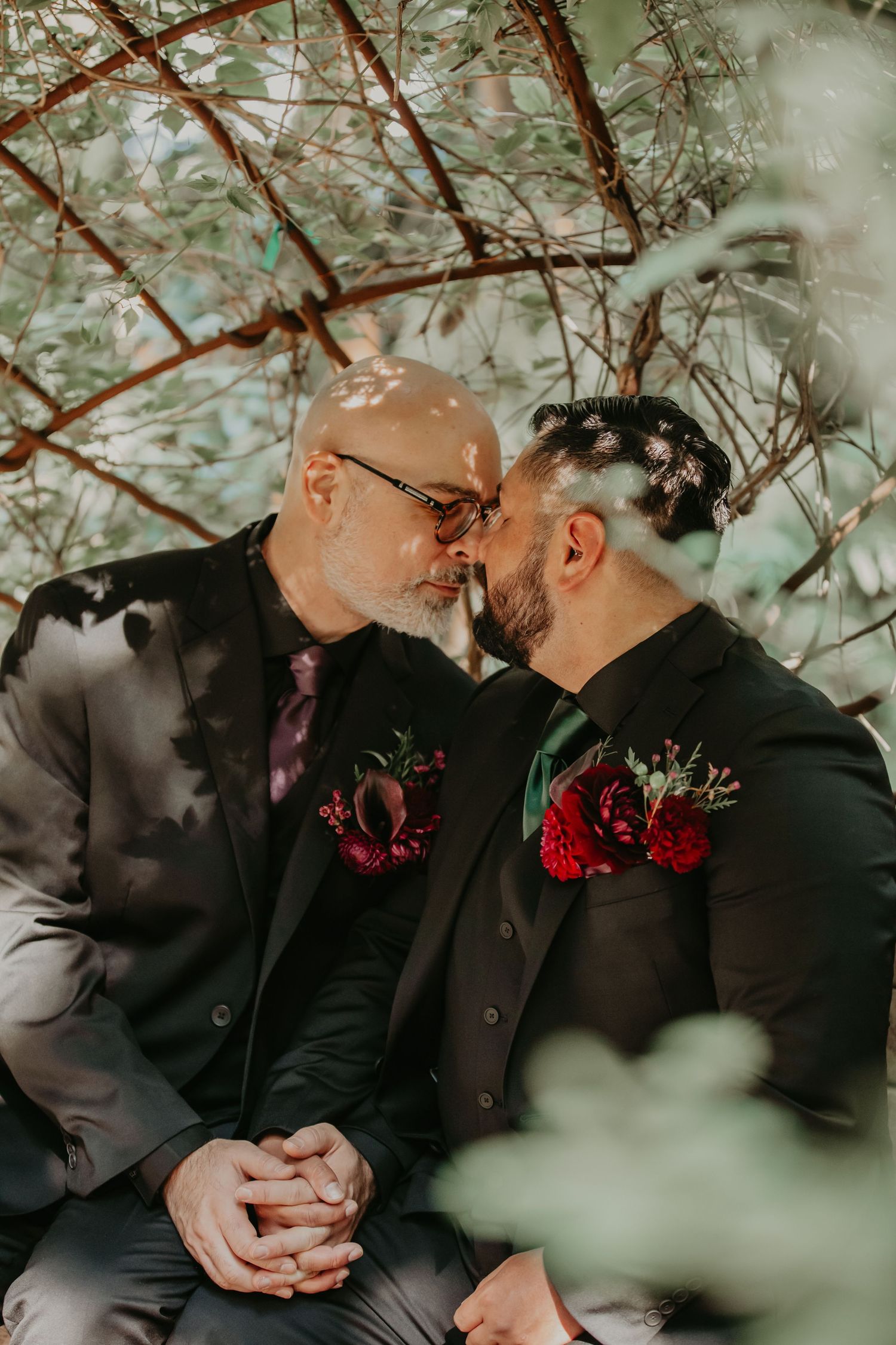 A romantic moment captured between two people wearing black suits with deep red boutonnieres in a garden setting.