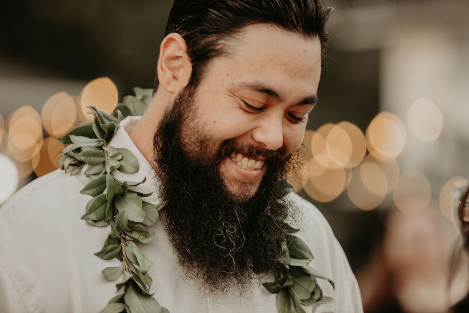 A smiling bearded person wearing a white dress shirt and leafy green lei garland around their neck with bokeh lights in background.