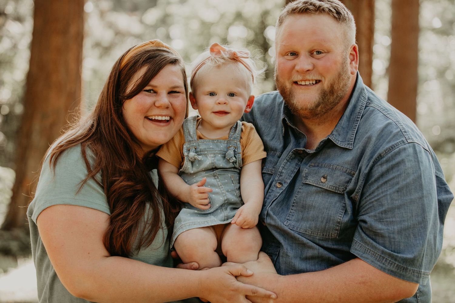 A family in casual attire poses together outdoors in a sunlit wooded setting.