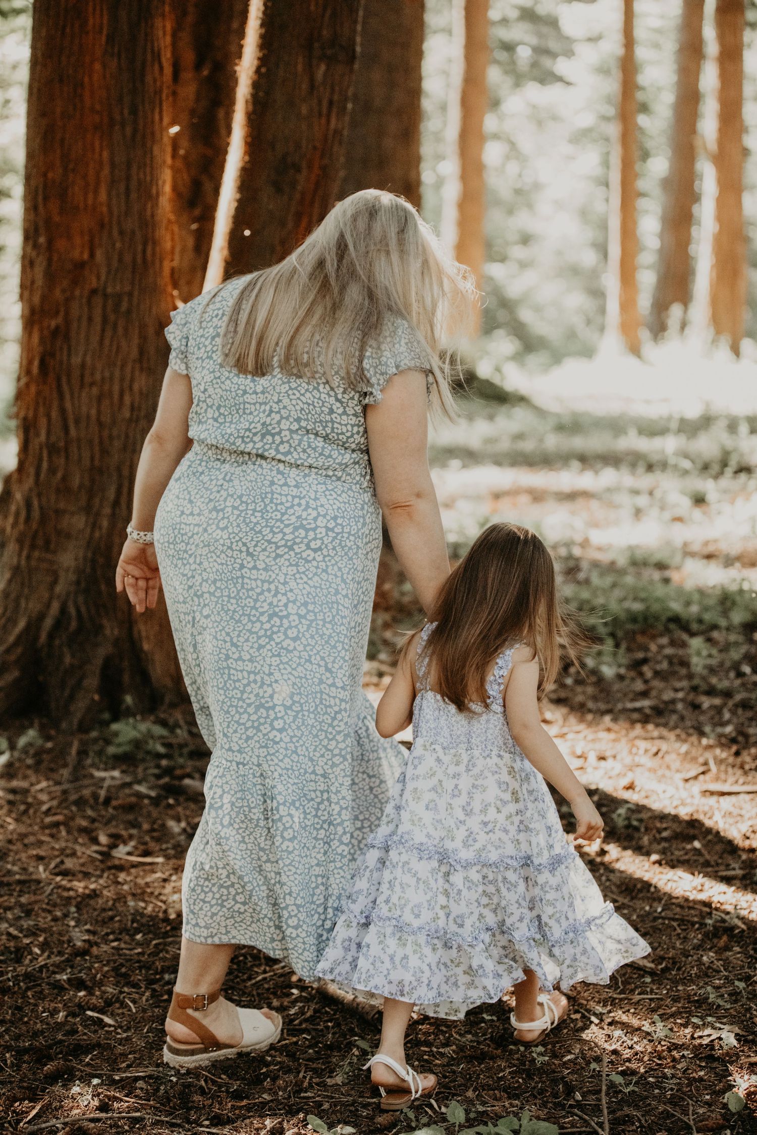 A mother and daughter wearing matching floral dresses walk hand in hand through a sunlit forest of tall trees.