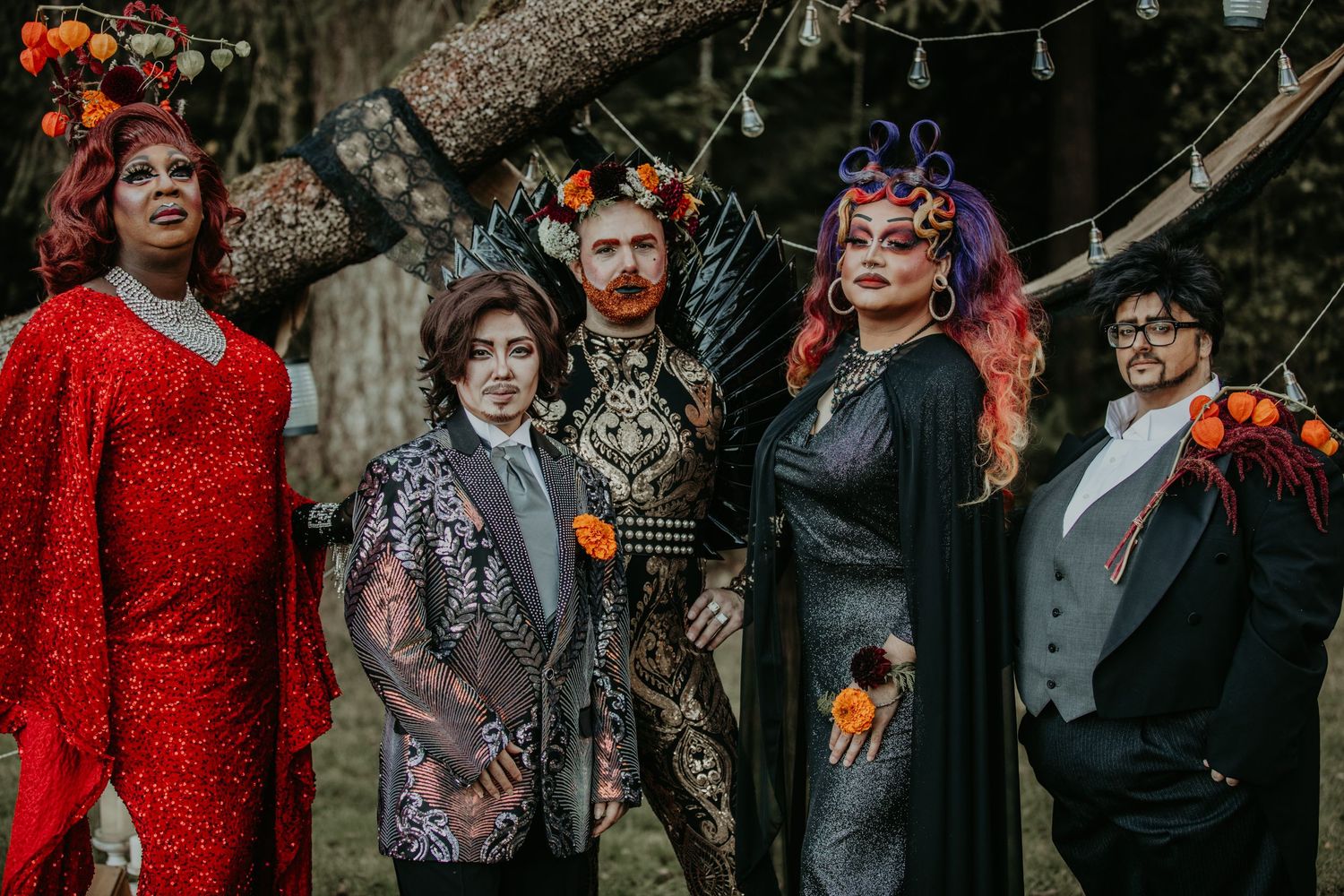 A group dressed in dark gothic Halloween attire with orange floral accents poses outdoors for a spooky autumn portrait.
