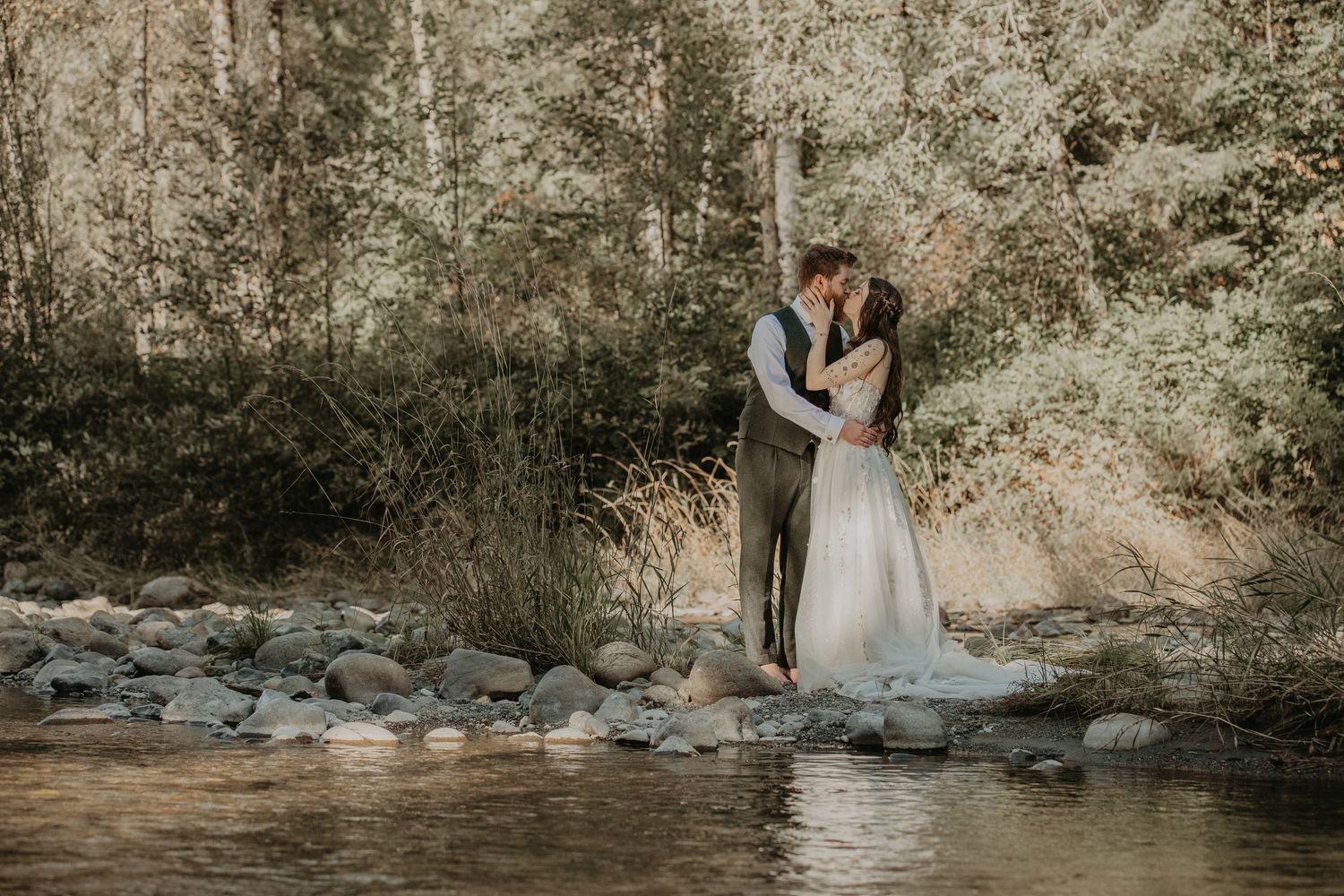A couple in wedding attire embraces near a stream in a sunlit forest setting.