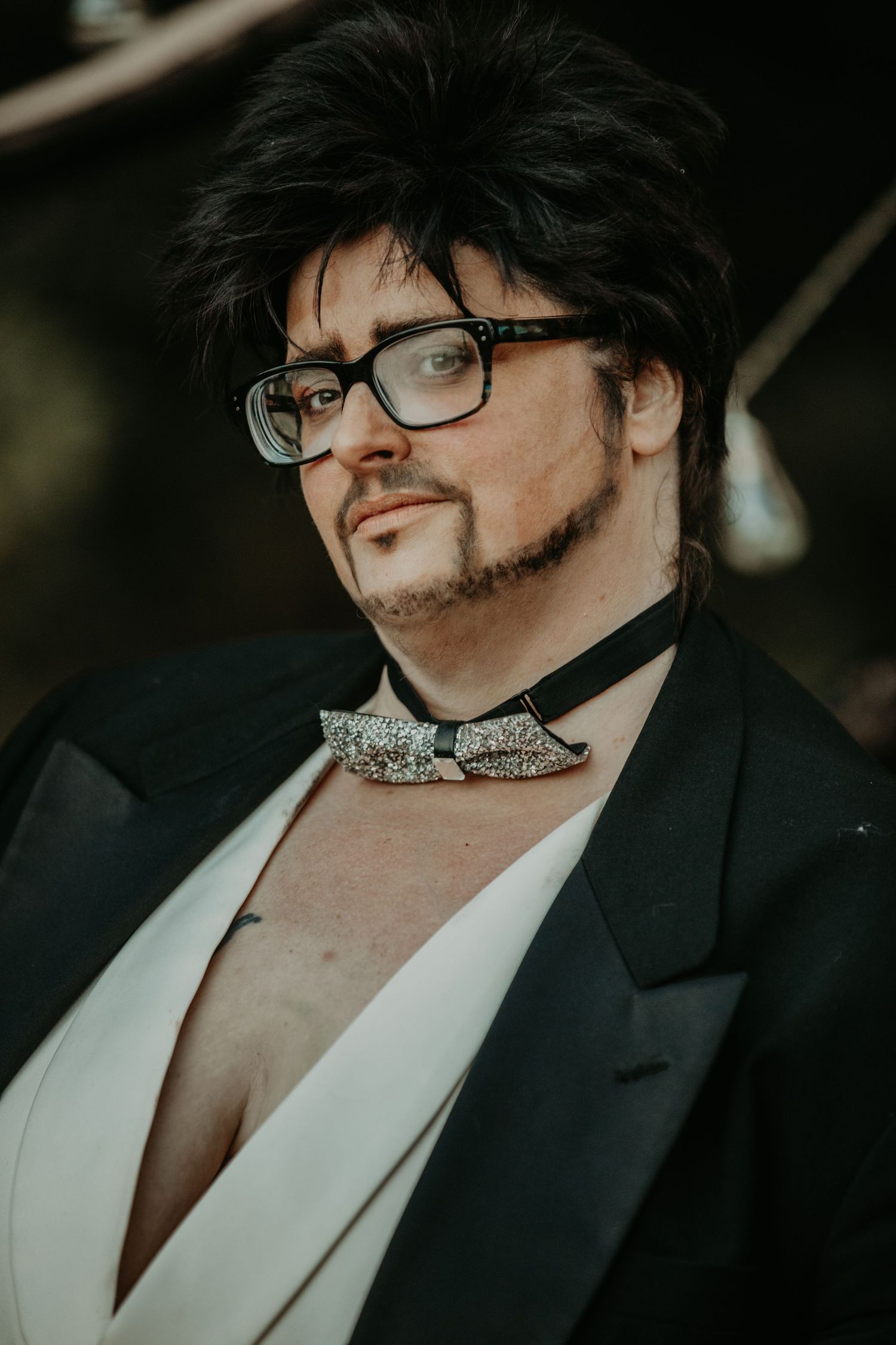 Close-up portrait of someone wearing glasses, black suit jacket, white shirt and sparkly bow tie in moody lighting.