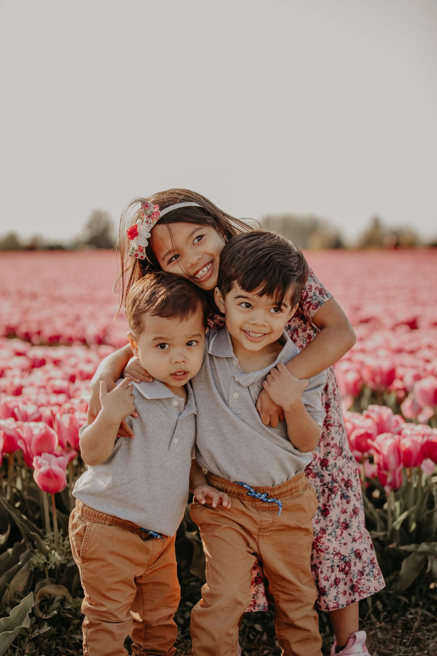 Three young siblings pose together in a vibrant pink tulip field during golden hour.