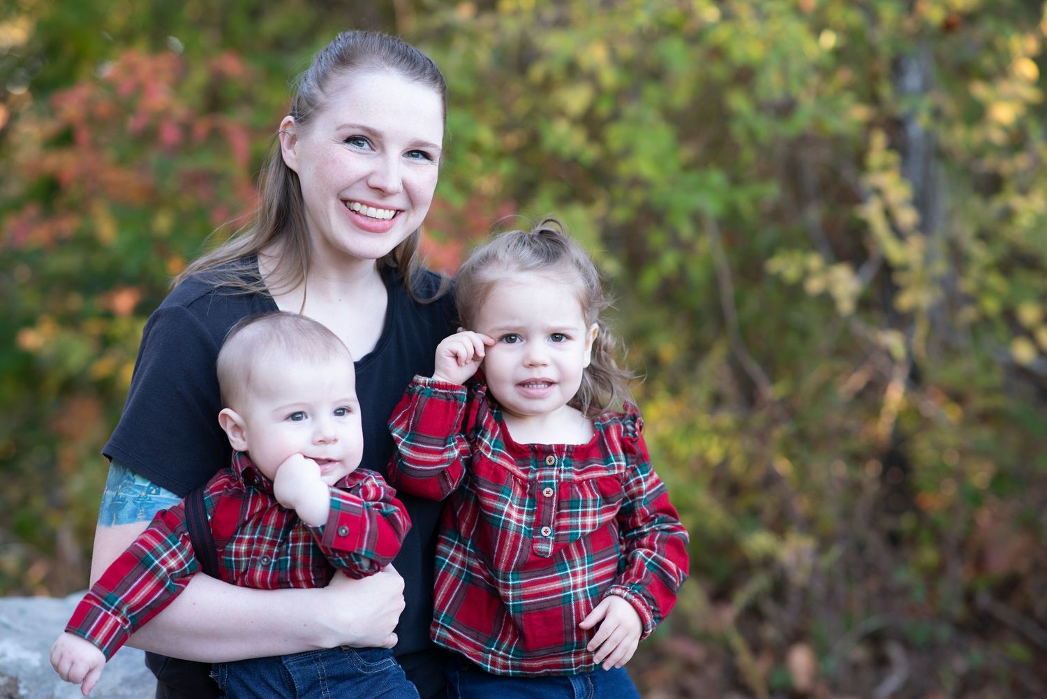 Family wearing coordinated red plaid and black outfits poses for outdoor autumn portraits in natural setting.