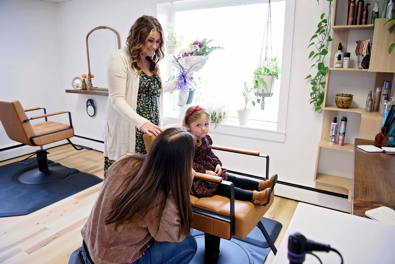 Three people gather in a bright room with bookshelves and plants, two seated and one standing during a salon session.