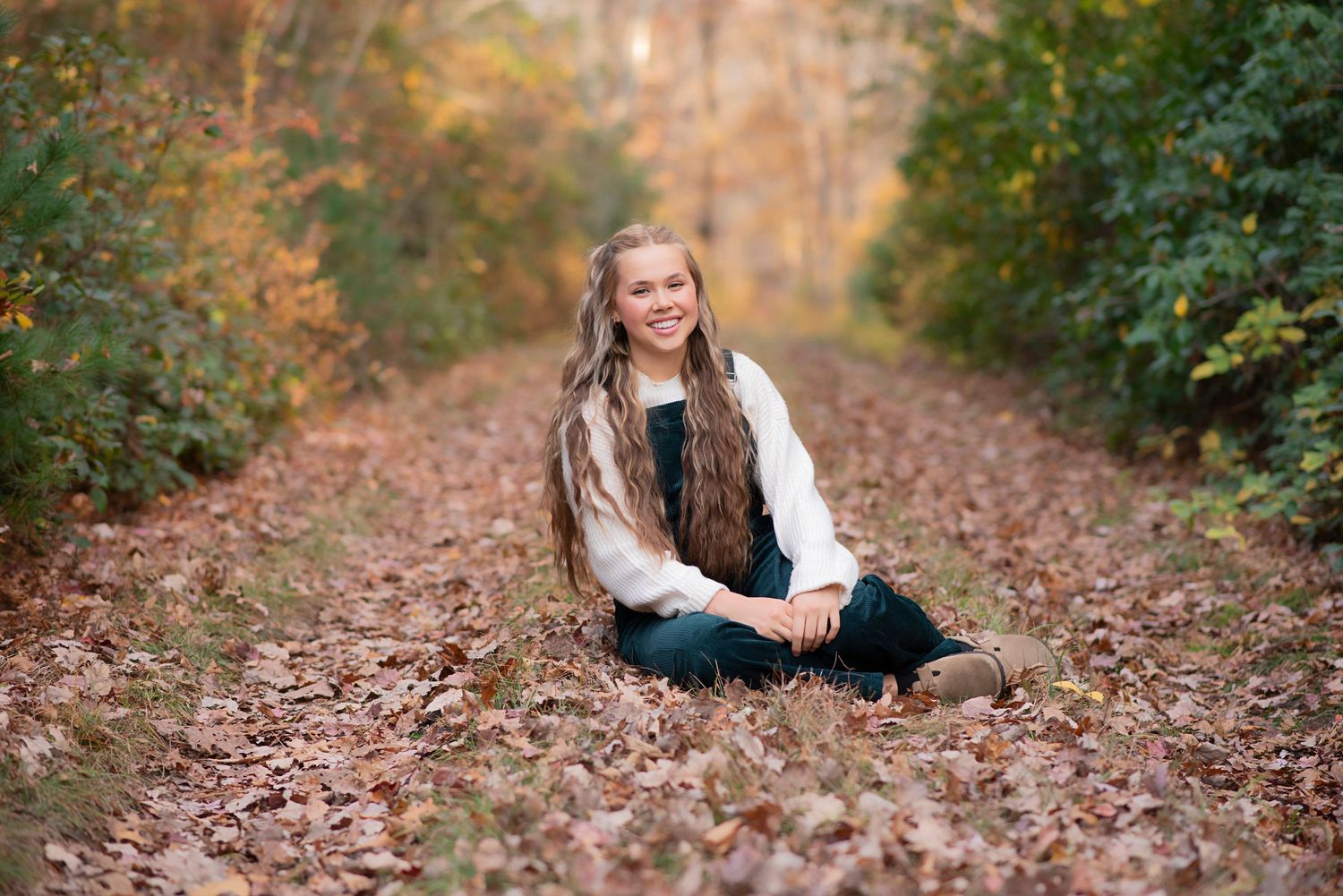 Autumn portrait on forest path covered in fallen leaves showing someone sitting on ground in casual sweater and jeans.