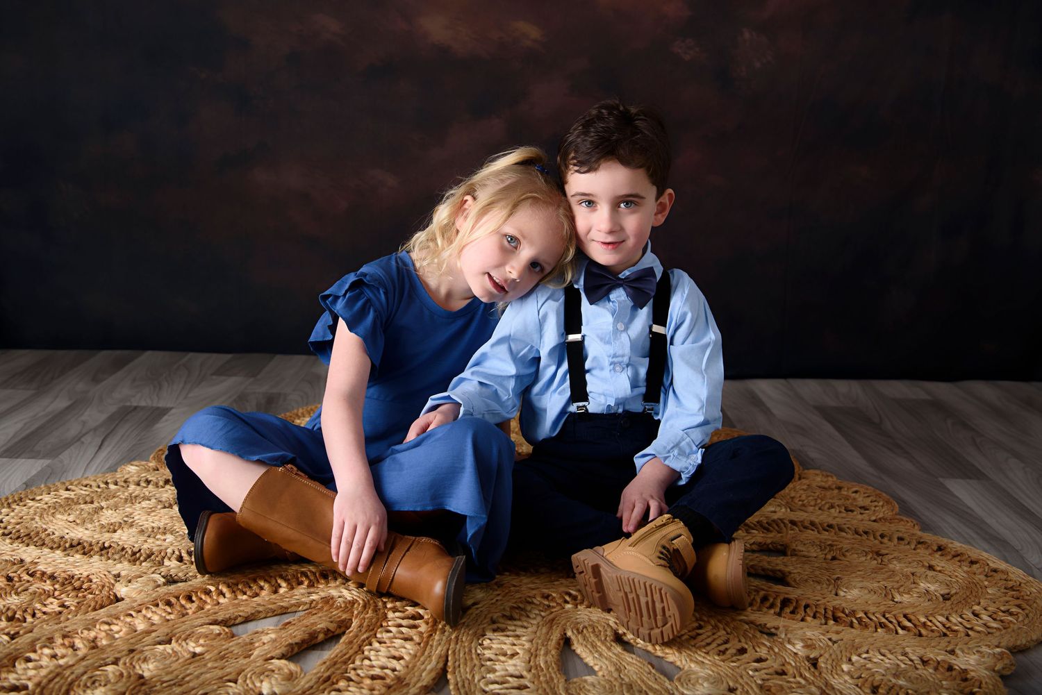 Two children in formal attire sit together on a brown rug against a dark backdrop in a studio setting.