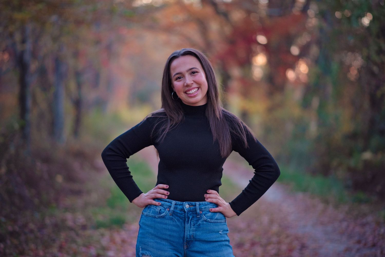 Someone wearing a black turtleneck and blue jeans poses on a forest path against a blurred autumn background.
