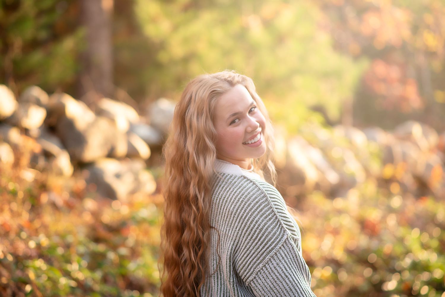 A woman with long wavy hair smiles over her shoulder while wearing a cozy grey sweater in autumn sunlight. Rhode Island Photographer