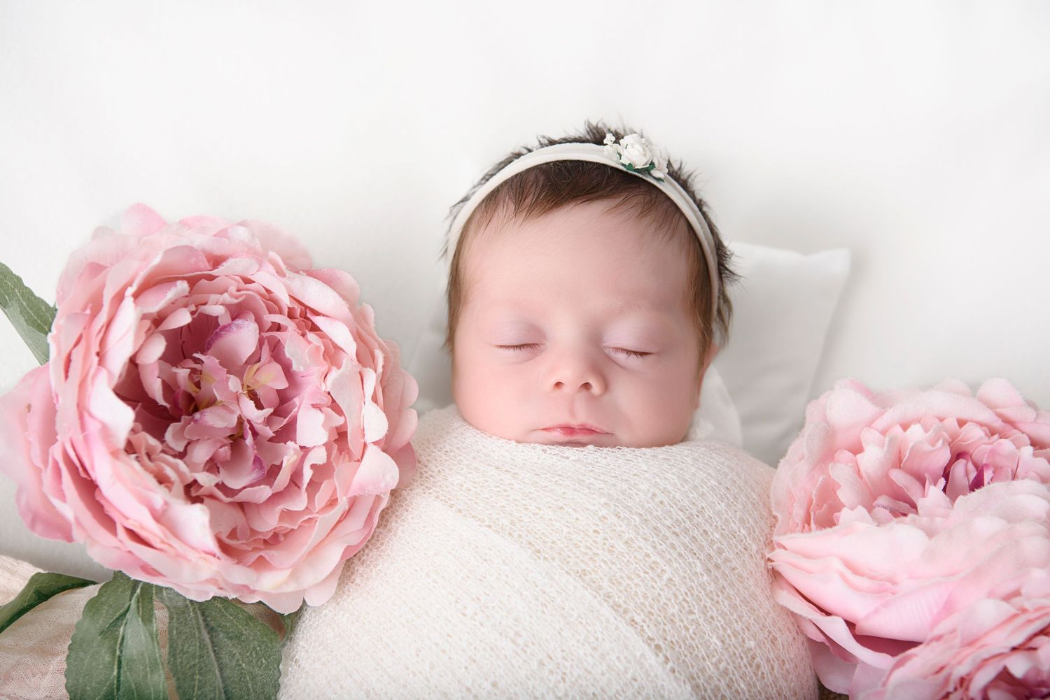 A sleeping newborn wearing a silver crown is swaddled in white fabric between two large pink peonies.
