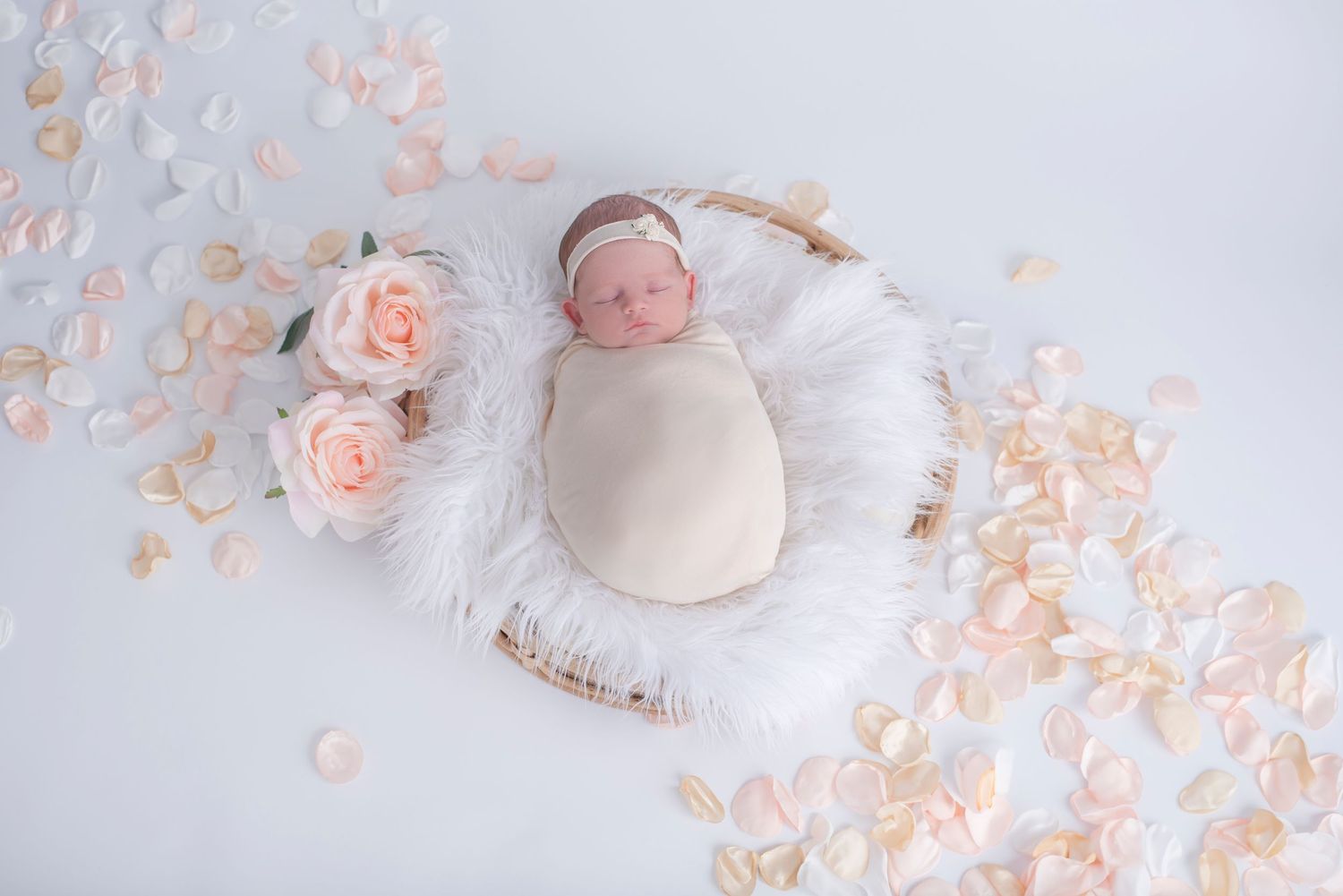 Newborn swaddled in white fabric lies on soft fur blanket surrounded by delicate pink rose petals and flowers.