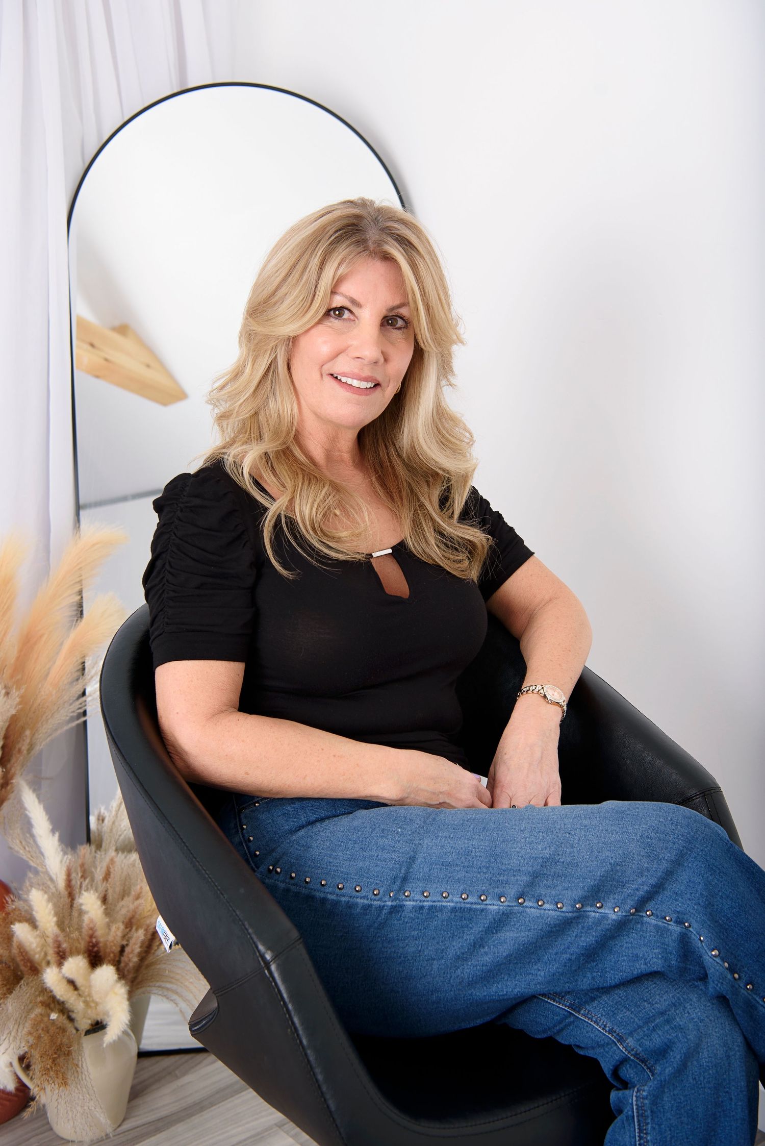 Person in black top and jeans lounging casually on a chair against white background.