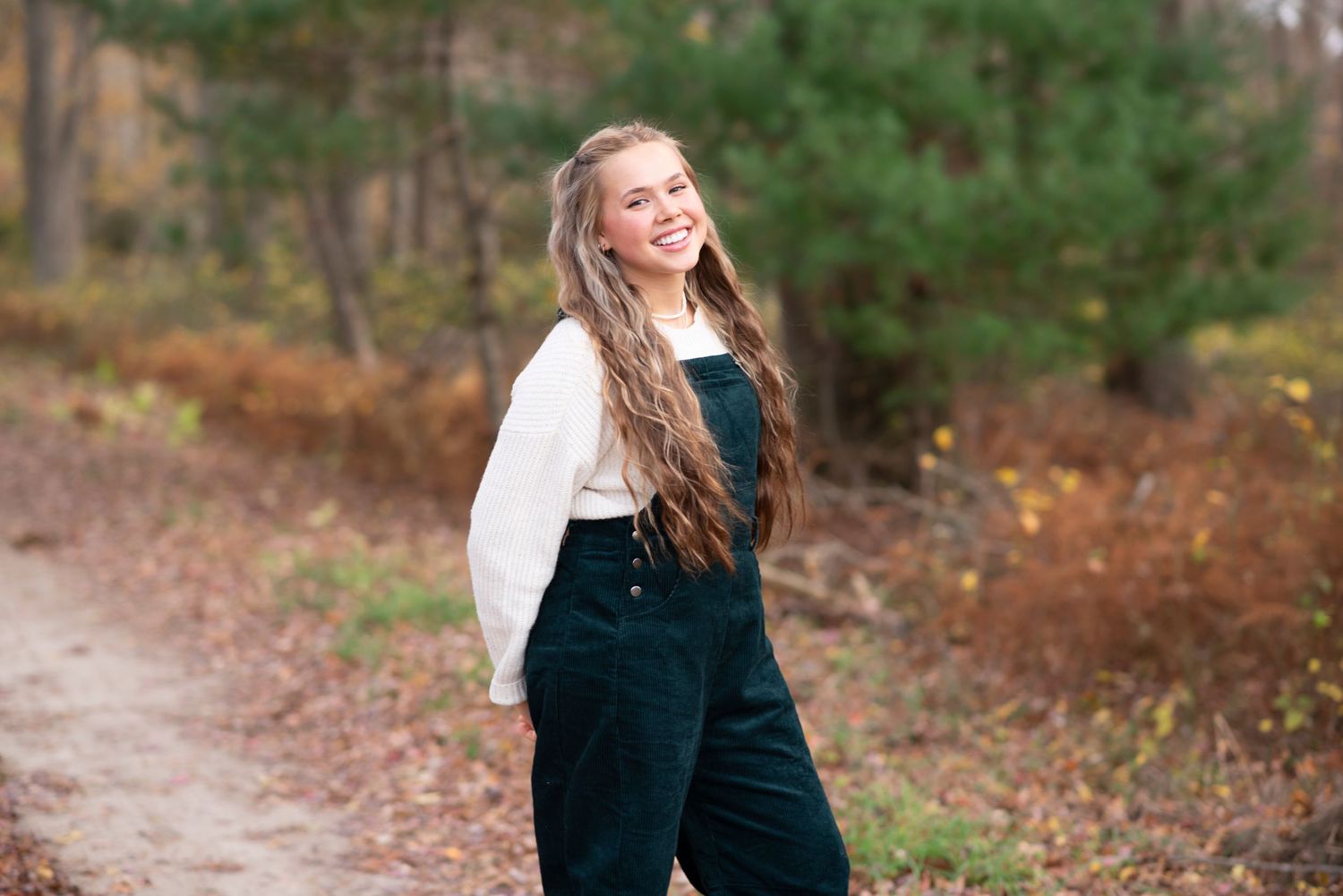 Someone wearing dark green corduroy overalls and a white sweater stands on an autumn forest path while smiling. Rhode Island Photographer