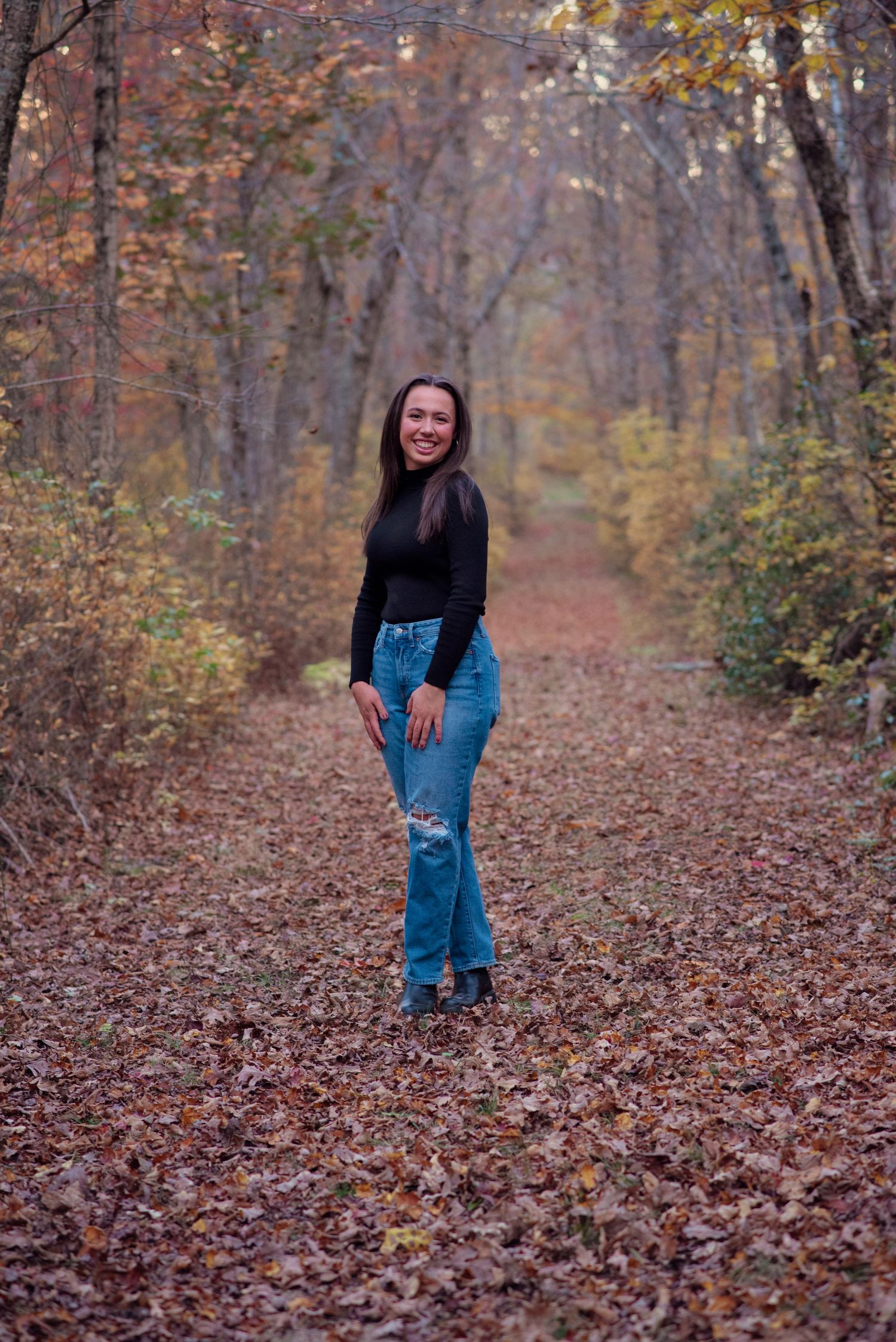 Someone in a black sweater and ripped jeans poses on an autumn forest path covered in fallen leaves.