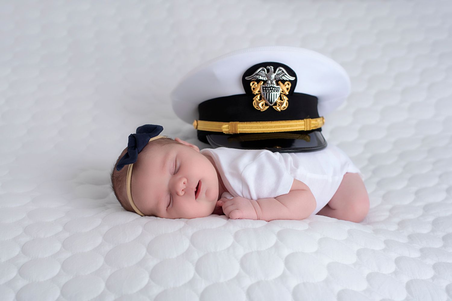 A newborn baby in a navy sailor uniform hat sleeps peacefully on a white blanket during a photography session.