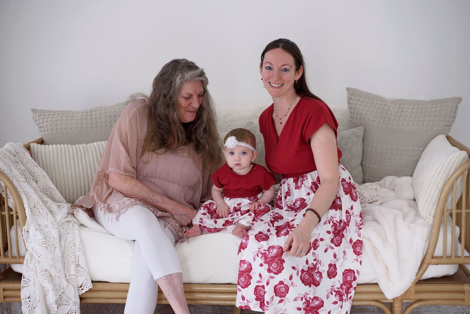 Three generations sit together on a cozy couch with white pillows and rose printed skirt.