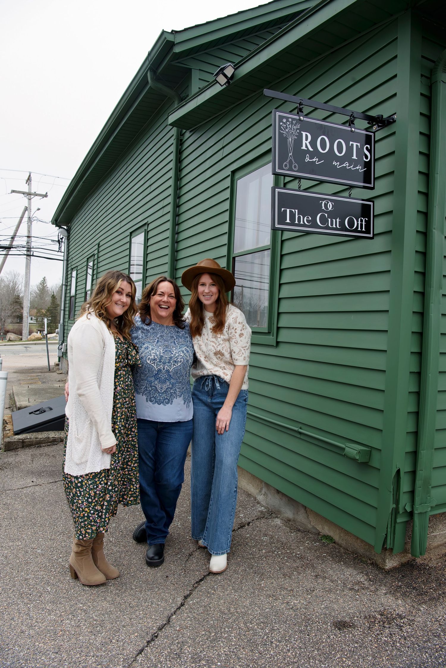 Three people stand together outside a green building with a 'Roots' and 'The Cut Off' sign mounted on the wall.