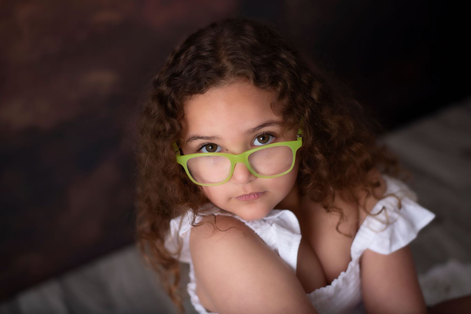 A young child with curly hair and green glasses smiles while wearing a white outfit.