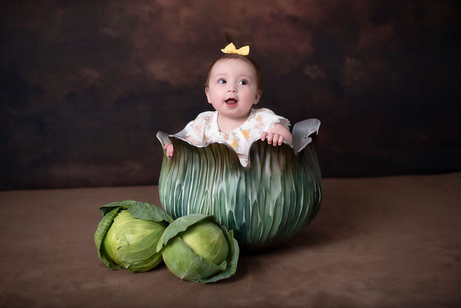 A baby wearing a small yellow bow sits inside a large decorative cabbage prop with real cabbages on the side.