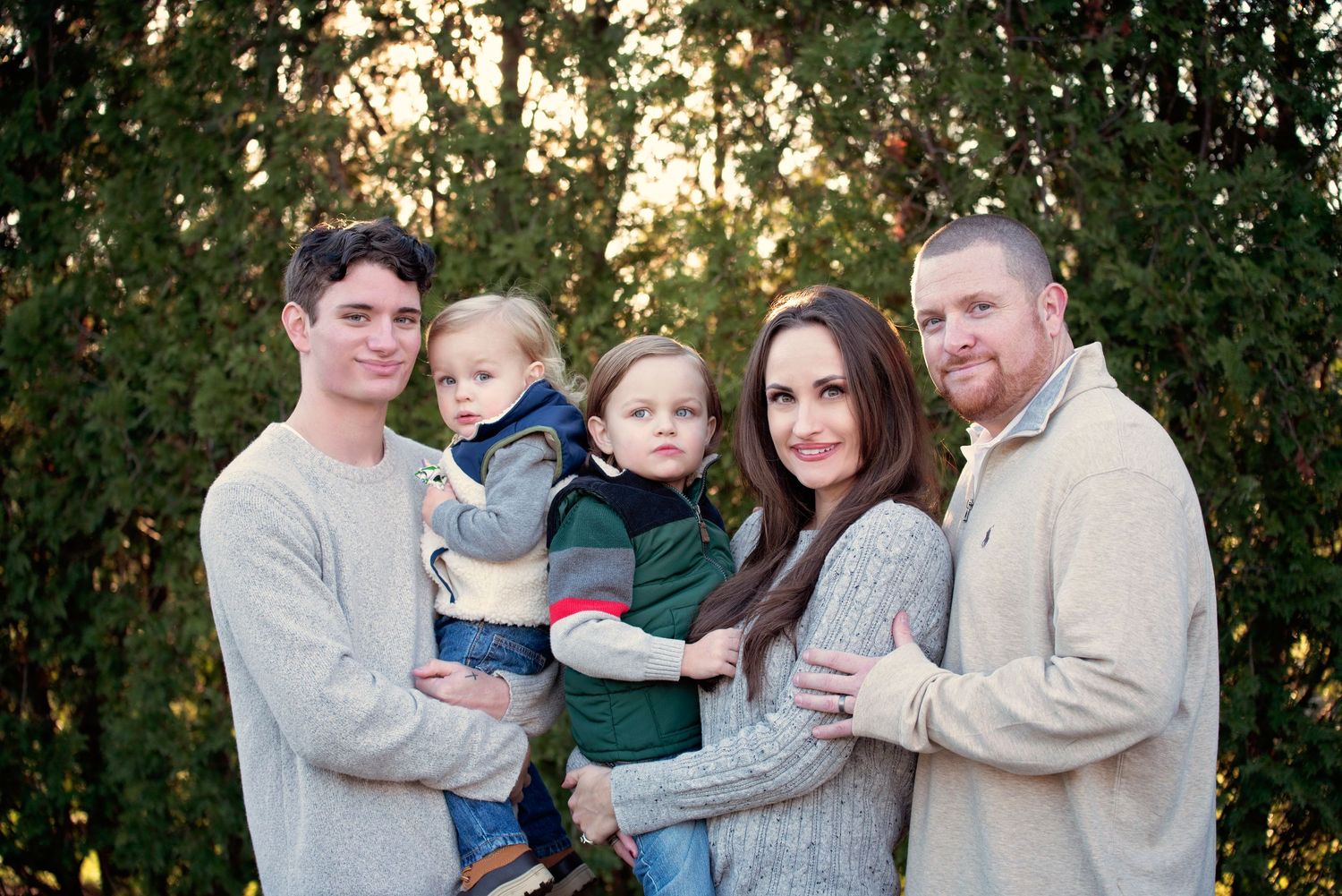 A happy family of five poses together outdoors against a leafy green background during sunset.