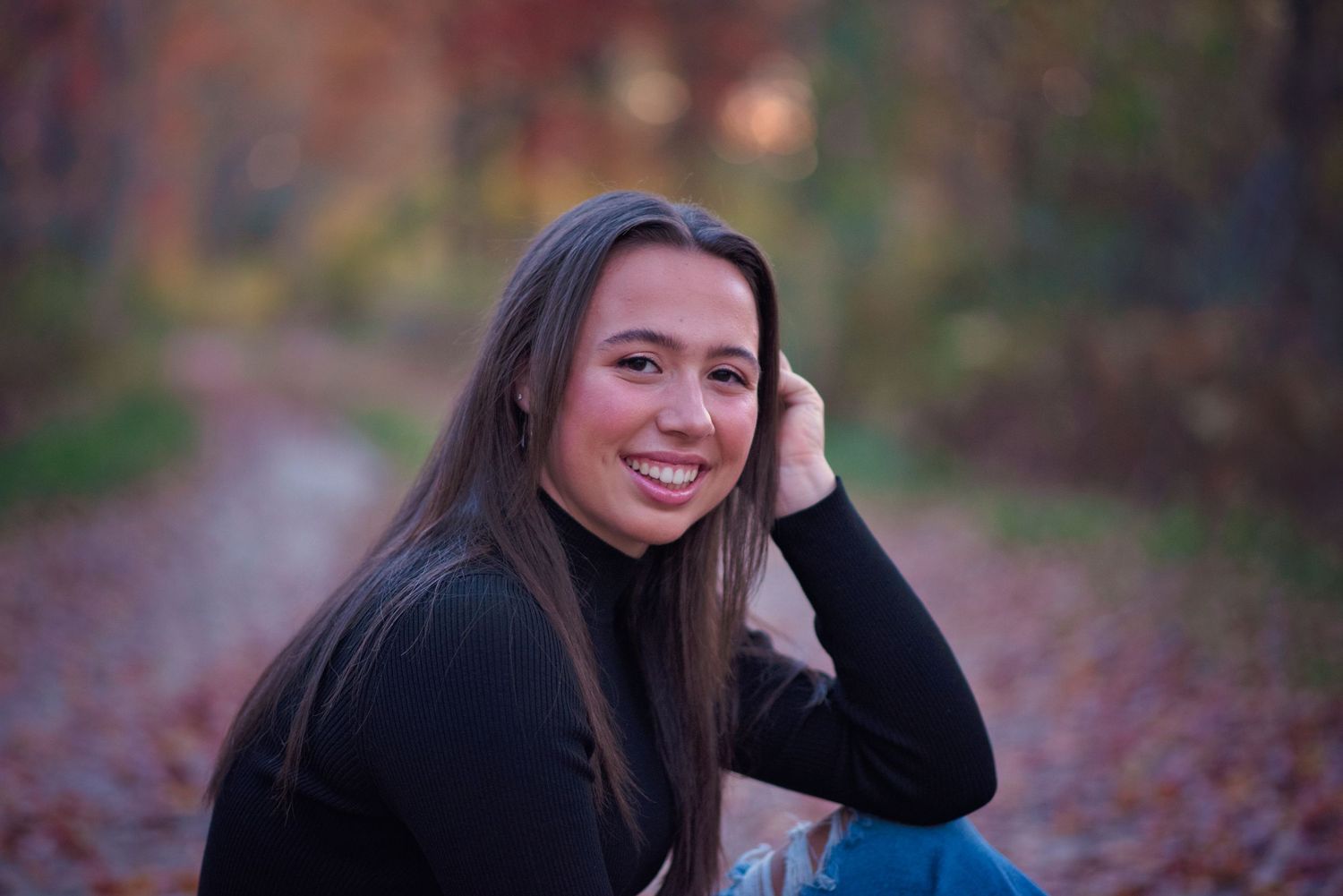 Someone seated outdoors on an autumn path with fallen leaves, wearing a black sweater and long dark hair.