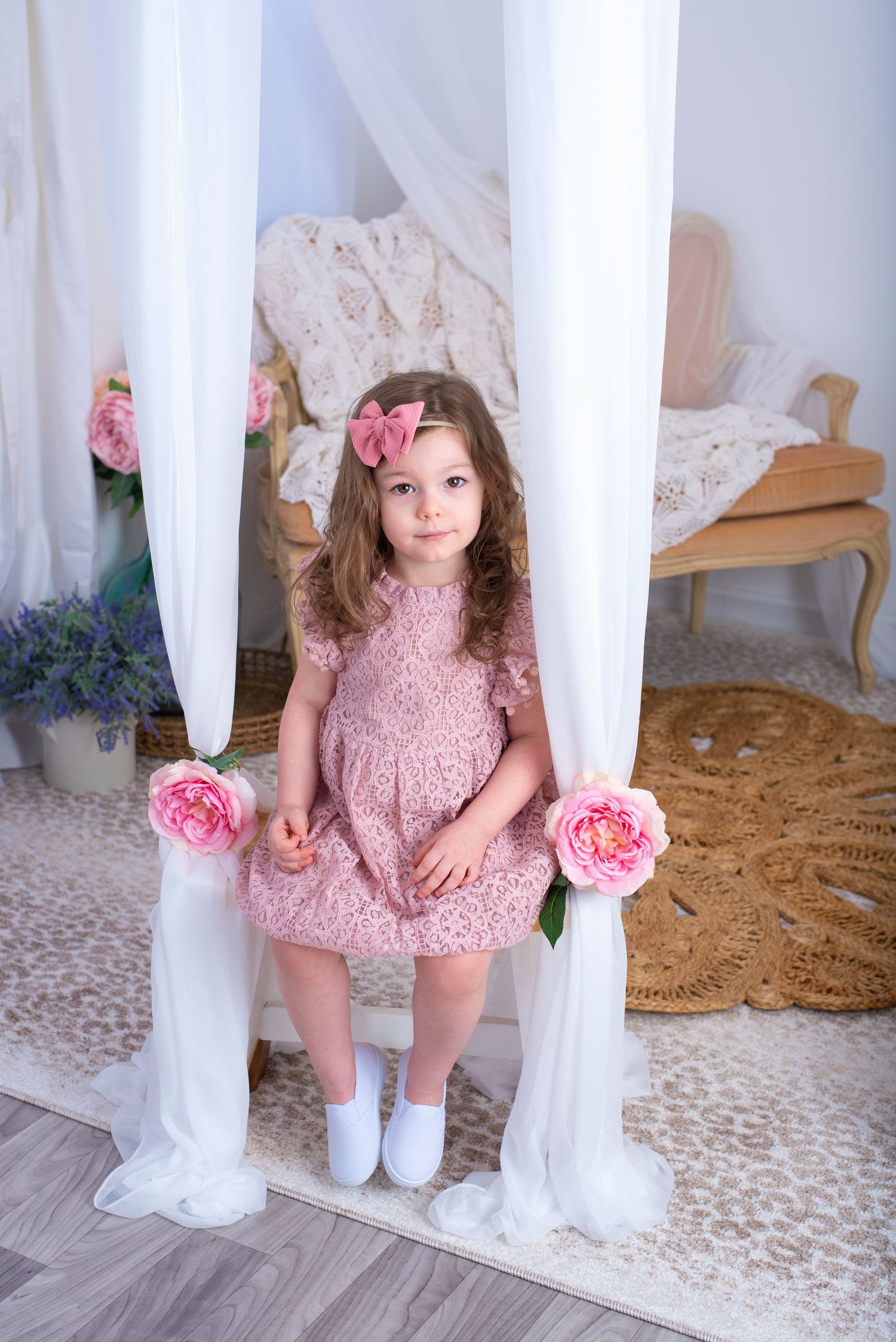 Small child in pink lace dress poses between white curtains decorated with pink roses in a romantic setting.