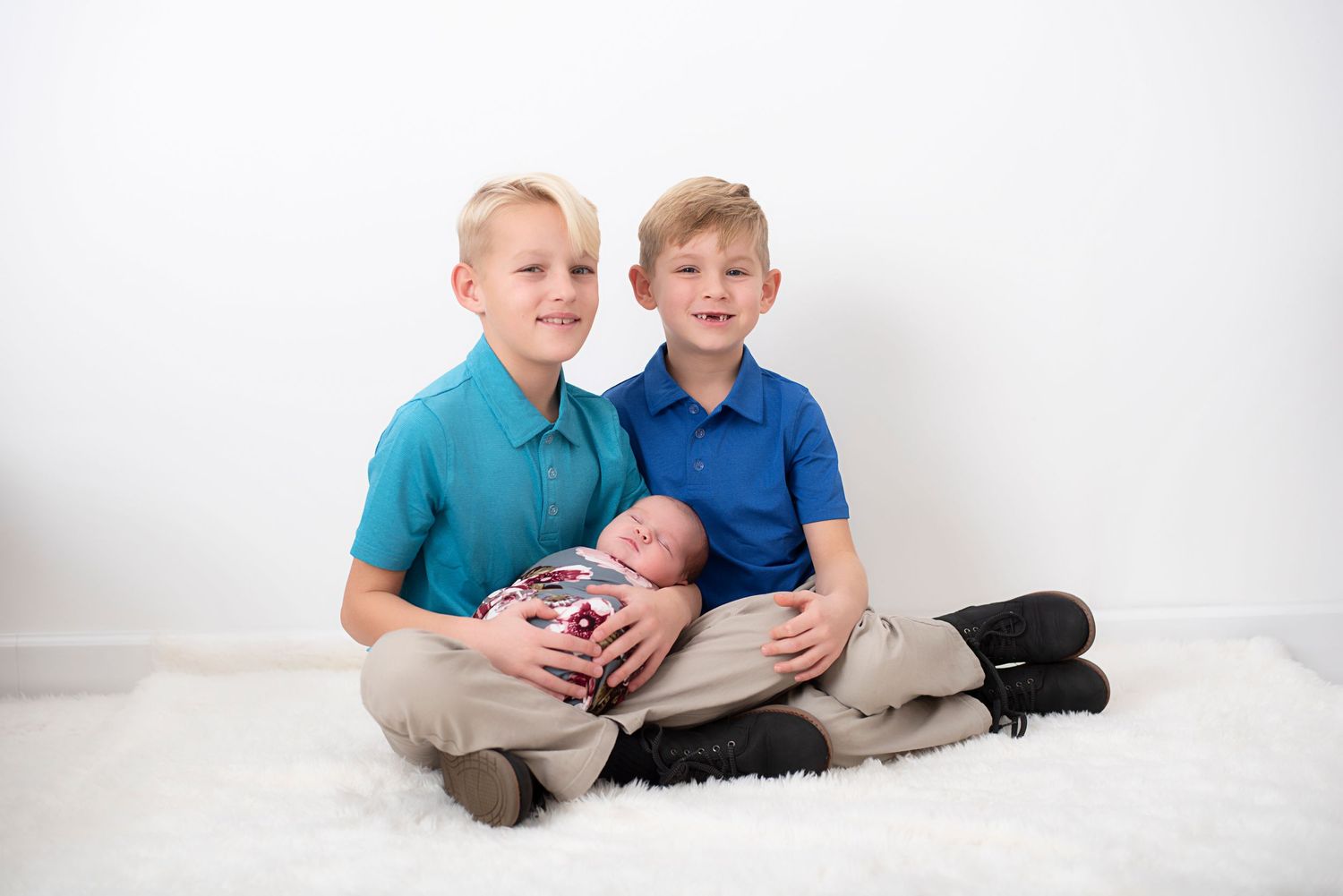 Two young siblings in polo shirts sitting on a white rug while holding their newborn baby sibling. Rhode Island Photographer
