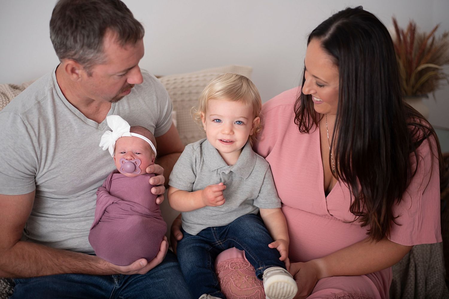 A family sits together on a couch with their young child and newborn baby in a warm indoor setting.