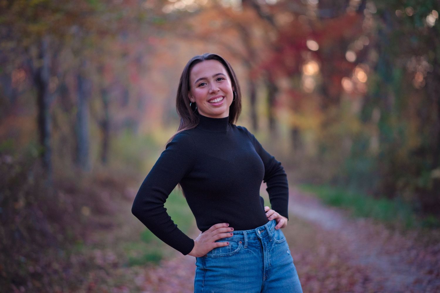 A person in a black sweater stands on an autumn forest trail surrounded by colorful fall foliage.
